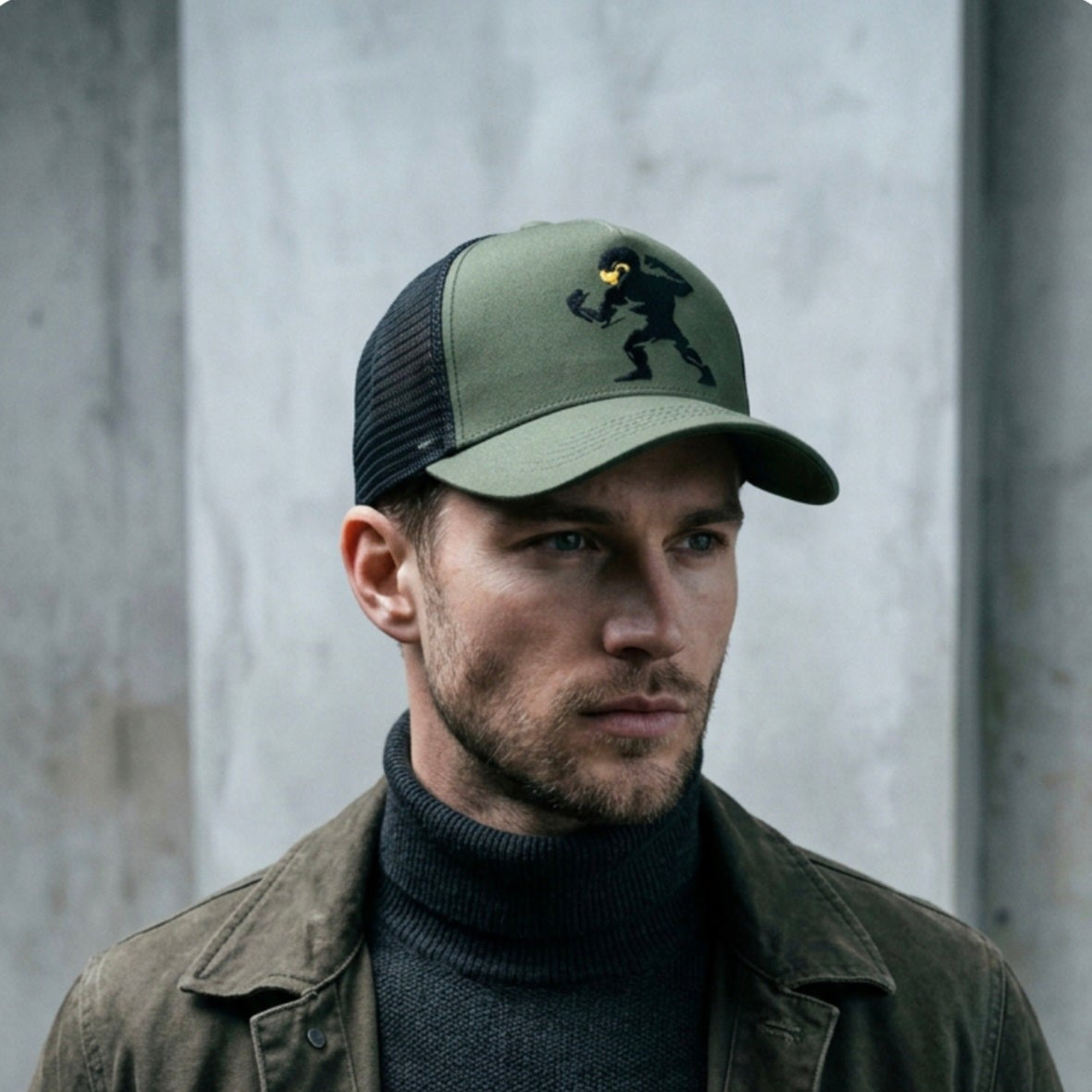 Man wearing a green cap  dooks nuts cap and dark jacket against a gray concrete wall.